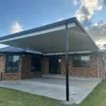 Flat insulated flyover patio roof with charcoal posts at Yugar attached to a brick home, over a concrete area beside a lawn and rural bushland