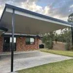 Flat insulated flyover patio roof with charcoal posts at Yugar attached to a brick home, over a concrete area beside a lawn and rural bushland