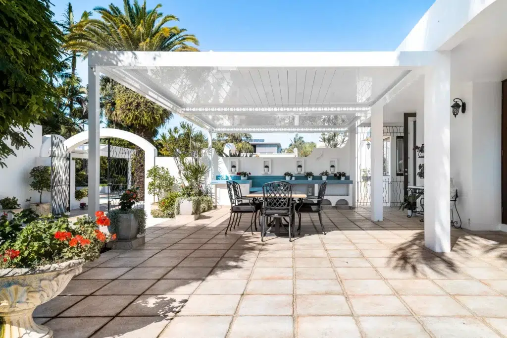 White louvred pergola patio with open louvres showing blue sky, outdoor dining set and red flower garden, Queensland