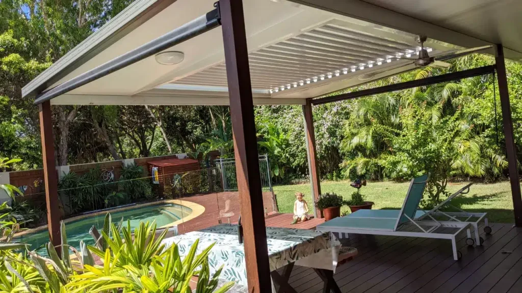 Combined insulated and louvred opening roof with timber-clad posts, LED strip lighting and ceiling fan at Wurtulla, Queensland over a timber deck, beside a swimming pool, lawn and tropical garden